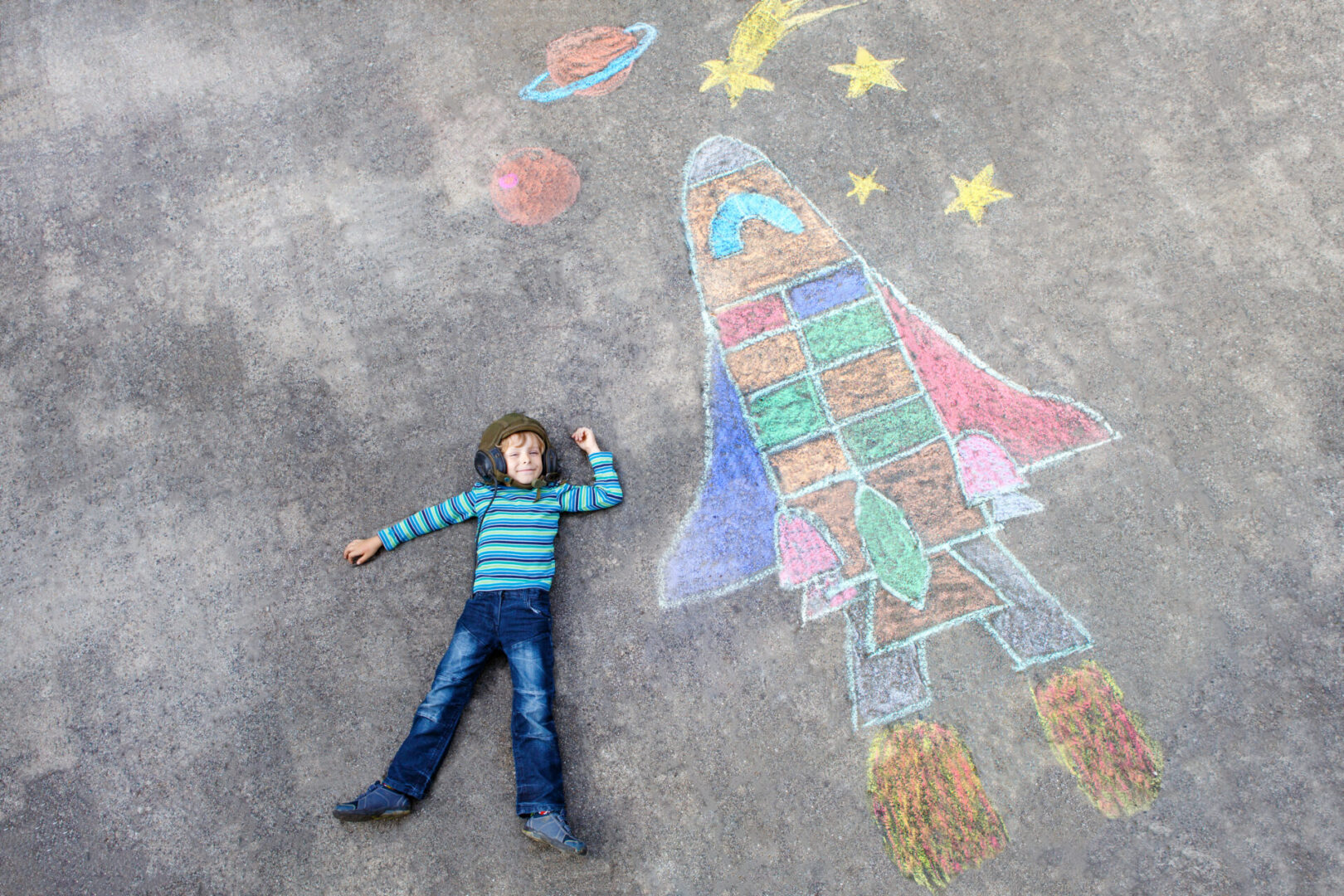 Funny little kid boy flying in universe by a space shuttle picture painting with colorful chalks. Creative leisure for children outdoors in summer.